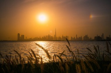Dubai 'nin altın saat manzaralı manzarası Burj Halife ile birlikte, suyun karşısında doğal ön planda çimlerle görülüyor. Şehir mimarisi ve huzurlu doğanın mükemmel bir karışımı. Dubai Creek Liman Manzarası.