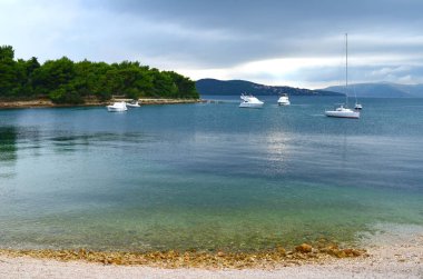 Hırvatistan 'ın Trogir kenti yakınlarındaki bir körfezin sakin sularında demirlemiş teknelerin huzurlu manzarası, çakıllı bir kıyı şeridi, ormanlık bir sahil ve bulutlu bir hava yaratıyor..