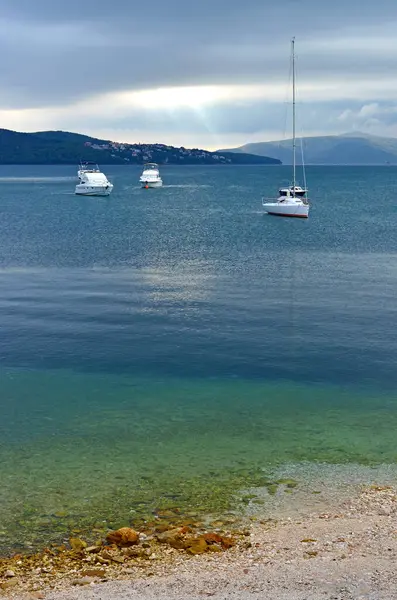 Hırvatistan 'ın Trogir kenti yakınlarındaki bir körfezin sakin sularında demirlemiş teknelerin huzurlu manzarası, çakıllı bir kıyı şeridi, ormanlık bir sahil ve bulutlu bir hava yaratıyor..