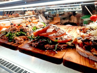 Ton balığı, domates, somon, peynir ve tohum gibi malzemelerle kaplı çeşitli açık sandviçler, iyi aydınlatılmış bir restoran vitrininde ahşap tahtalarda sergileniyor..