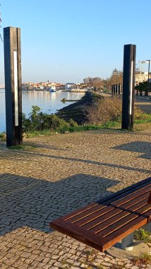 Seixal gezinti alanında sakin bir şafak sahnesi. Nehir kenarındaki ağaçların kaldırımlı boş bir bank ve suyun karşısındaki zıt binaların huzurlu yansıması var.
