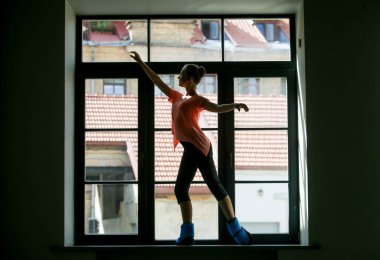 Silhouette of a young ballerina on the window sill, roof in the background. Concept of healthy lifestyle