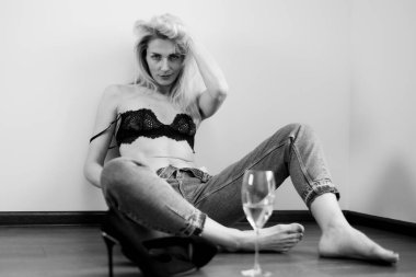 Kot pantolonlu bir kız, siyah bir sutyen ve beyaz bir bardak şarap yerde oturuyor.