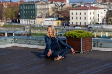 Mavi ceketli, koyu renk gömlekli ve kot pantolonlu güzel bir kadın Krakow şehrinin arka planında poz veriyor.