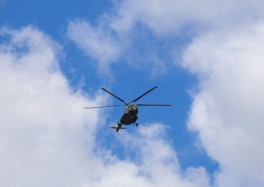 Mavi gökyüzünde bulutlarla uçan bir helikopterin silüeti.