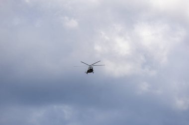 Mavi gökyüzünde bulutlarla uçan bir helikopterin silüeti.
