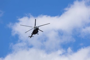 Mavi gökyüzünde bulutlarla uçan bir helikopterin silüeti.