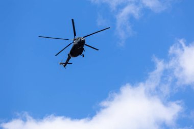 Mavi gökyüzünde bulutlarla uçan bir helikopterin silüeti.
