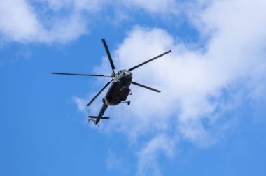 Mavi gökyüzünde bulutlarla uçan bir helikopterin silüeti.