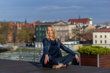 Mavi ceketli, koyu renk gömlekli ve kot pantolonlu güzel bir kadın Krakow şehrinin arka planında poz veriyor.