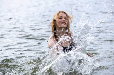 Neşeli kız kameraya su sıçratıyor. Sıçramaya odaklan. Kumsaldaki güzel genç kadın elleriyle su sıçratıyor.