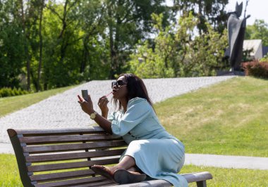 Güzel, koyu tenli bir kız huzurlu bir ortamda dolgun dudaklarına ruj sürüyor. Parktaki bankta bir güzellik anı.