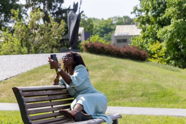 Güzel, koyu tenli bir kız huzurlu bir ortamda dolgun dudaklarına ruj sürüyor. Parktaki bankta bir güzellik anı.