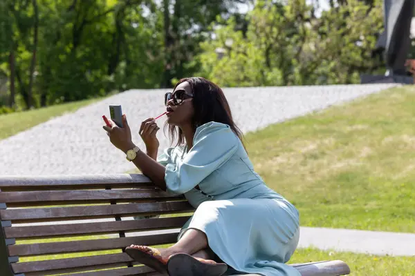 Güzel, koyu tenli bir kız huzurlu bir ortamda dolgun dudaklarına ruj sürüyor. Parktaki bankta bir güzellik anı.