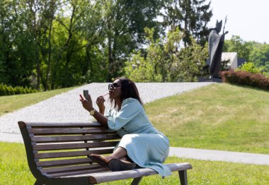 Güzel, koyu tenli bir kız huzurlu bir ortamda dolgun dudaklarına ruj sürüyor. Parktaki bankta bir güzellik anı.