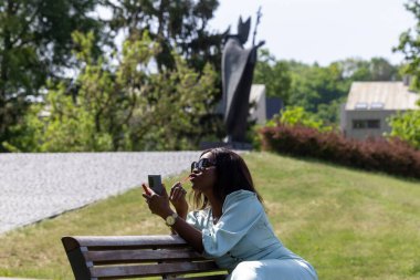 Güzel, koyu tenli bir kız huzurlu bir ortamda dolgun dudaklarına ruj sürüyor. Parktaki bankta bir güzellik anı.