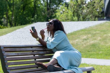 Güzel, koyu tenli bir kız huzurlu bir ortamda dolgun dudaklarına ruj sürüyor. Parktaki bankta bir güzellik anı.