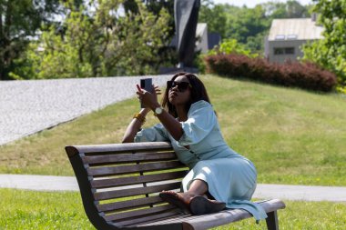 Güzel, koyu tenli bir kız huzurlu bir ortamda dolgun dudaklarına ruj sürüyor. Parktaki bankta bir güzellik anı.