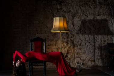 Kırmızı elbiseli zarif bir kadın, eski moda bir dekor içinde, lambanın altında poz veriyor.