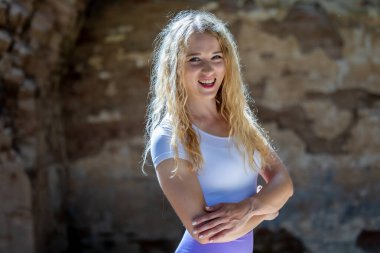 Beyaz gömlekli sarışın bir kadın, güneşli bir ortamda gülümsüyor, bulanık bir arka planda doğal güzellik yayıyor.