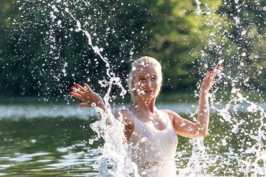 Beyaz bir yaz elbisesi içinde neşeli genç bir kadın güneşli bir günde doğal bir gölde su sıçratarak eğleniyor. Köpüklü su damlalarıyla havada yakalanan enerji hareketi doğada özgürlük, tazelik ve mutluluk hissi yaratıyor. Kişi başı