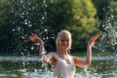 Beyaz bir yaz elbisesi içinde neşeli genç bir kadın güneşli bir günde doğal bir gölde su sıçratarak eğleniyor. Köpüklü su damlalarıyla havada yakalanan enerji hareketi doğada özgürlük, tazelik ve mutluluk hissi yaratıyor. Kişi başı