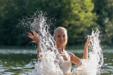 Beyaz bir yaz elbisesi içinde neşeli genç bir kadın güneşli bir günde doğal bir gölde su sıçratarak eğleniyor. Köpüklü su damlalarıyla havada yakalanan enerji hareketi doğada özgürlük, tazelik ve mutluluk hissi yaratıyor. Kişi başı
