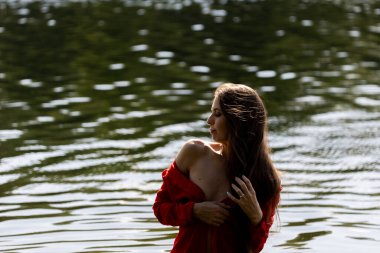 Dalgalı kırmızı elbiseli zarif genç bir kadın gölün kenarında poz veriyor. Sakin su sakin bir zemin oluştururken, doğal ışık onun zarif güzelliğini ve yumuşak ifadesini vurgular.