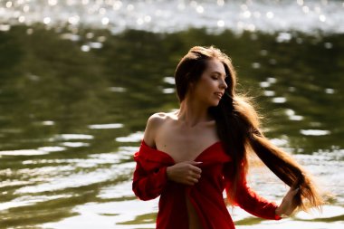 Dalgalı kırmızı elbiseli zarif genç bir kadın gölün kenarında poz veriyor. Sakin su sakin bir zemin oluştururken, doğal ışık onun zarif güzelliğini ve yumuşak ifadesini vurgular.