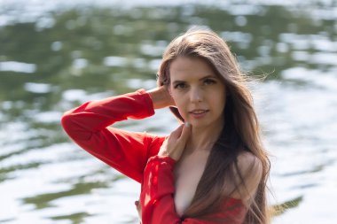 Dalgalı kırmızı elbiseli zarif genç bir kadın gölün kenarında poz veriyor. Sakin su sakin bir zemin oluştururken, doğal ışık onun zarif güzelliğini ve yumuşak ifadesini vurgular.