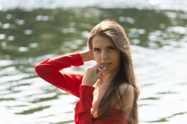 Dalgalı kırmızı elbiseli zarif genç bir kadın gölün kenarında poz veriyor. Sakin su sakin bir zemin oluştururken, doğal ışık onun zarif güzelliğini ve yumuşak ifadesini vurgular.
