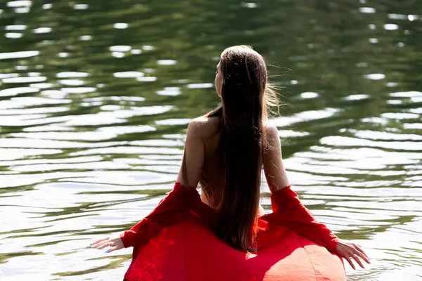 Uzun saçlı zarif bir genç kadın, arkadan görülüyor, sığ göl suyunda duruyor. Kırmızı elbisesi meltemde usulca süzülüyor, doğal ortamda sakin ve romantik bir hava yaratıyor..