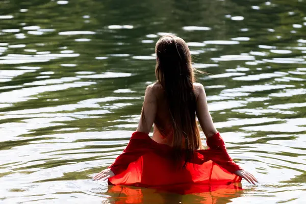 Uzun saçlı zarif bir genç kadın, arkadan görülüyor, sığ göl suyunda duruyor. Kırmızı elbisesi meltemde usulca süzülüyor, doğal ortamda sakin ve romantik bir hava yaratıyor..