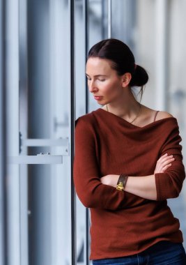 Calm thoughtful woman standing by the large window, wearing a brown sweater and jeans, looking outside with soft natural light reflection. Modern interior portrait