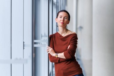 Calm thoughtful woman standing by the large window, wearing a brown sweater and jeans, looking outside with soft natural light reflection. Modern interior portrait