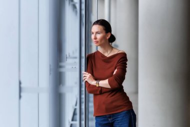 Calm thoughtful woman standing by the large window, wearing a brown sweater and jeans, looking outside with soft natural light reflection. Modern interior portrait