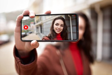 Sokaktaki genç bir kadın telefonunda video blogu yazıyor. Görüntüsü telefonun ekranında görünüyor..