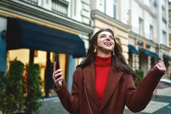Excited trendy woman celebrating good news holding phone in the evening street