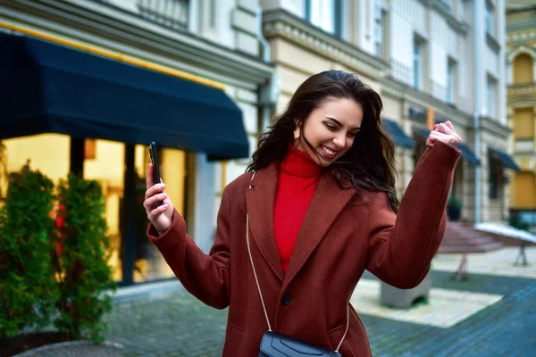 Beautiful Overjoyed young brunette woman with long hair in a red coat celebrating good news holding phone and clenching fist in the evening street