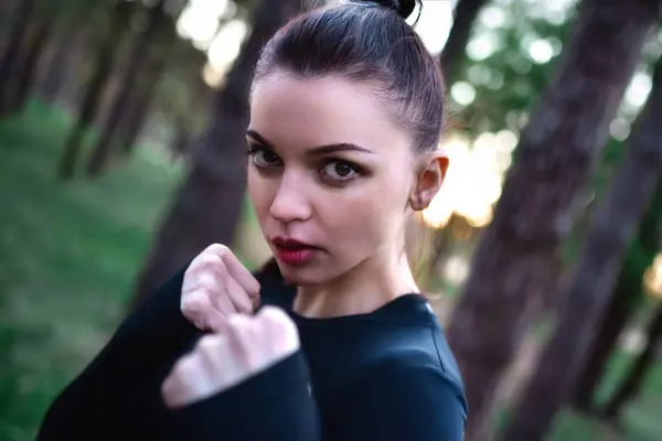 stock image Young woman doing martial arts training in sporty black top holding hands in fighting pose against forest background.