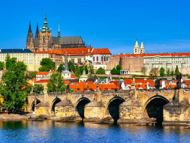 Çek Cumhuriyeti, Prag 'da açık mavi gökyüzü altında Charles Köprüsü ve tarihi Prag Kalesi' nin manzarası. Geleneksel kırmızı çatılar, Gotik mimari ve canlı yaz yeşilliği.
