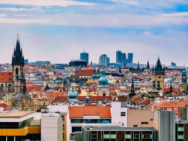 Prag 'ın ufuk çizgisinin canlı panoramik manzarası, tarihi kuleler ve modern mimarinin bir karışımını sergiliyor. Sahne, yumuşak mavi gökyüzünün altındaki terracotta çatıları arasındaki ikonik işaretleri vurguluyor.