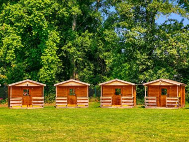 Lush Trees tarafından çevrelenmiş yeşil çimenlikte dört küçük tahta kulübe, Kamp, Glamping, Eco Retreats, Kırsal Turizm ve Doğa Yaşam Tarzı İçin İdeal