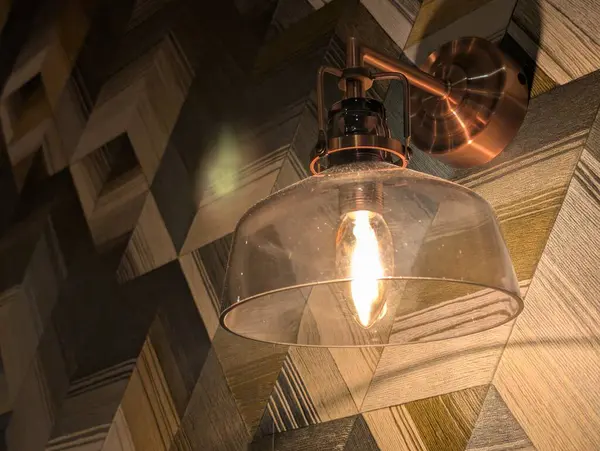 Temiz cam gölgeli bakır duvar lambası, geometrik ahşap arka plan, sıcak ışık. Yüksek kalite fotoğraf