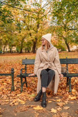 Havalı, güzel, sıcak bir sonbahar gününde, örülmüş bir şapkadan çıkan kalın bir paltoyla sonbahar kadını bir bankta dinlenir.