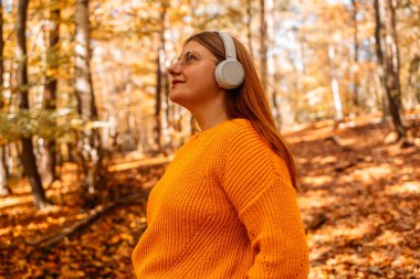Parlak, şık, sarı kazaklı gülümseyen bir kadın akıllı telefon kullanarak ve müzik dinleyerek harika bir ruh hali içinde. Kapalı gözlü kız sonbahar ormanında akıllı telefonuyla müzik listesinin keyfini çıkarıyor.