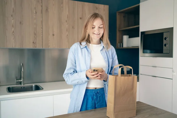 stock image Young lady unpacking paper bag after shopping using smartphone in kitchen interior at home, copy space.