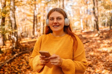 Parkta gülümseyen güzel beyaz bir kadının görüntüsü. Açık havada kulaklıkla müzik dinliyor. Cep telefonu kullanıyor..