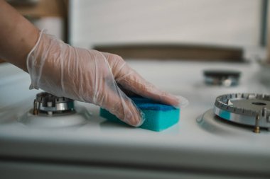 Mutfakta süngerle gaz sobası temizleyen kadın, yakın plan. Ocağın üstünü bulaşık süngeriyle temizlerken lastik eldiven takan ellerin yakın çekimi.
