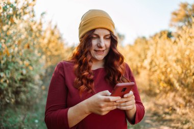 Neşeli gülümseyen ve cep telefonu kullanan mutlu, beyaz bir kadın, sonbahar tatilinde iyi vakit geçirir. Ekim günü dışarıda, kızıl saçlı ve gözlüklü sevimli bir hanımın açık hava portresi.